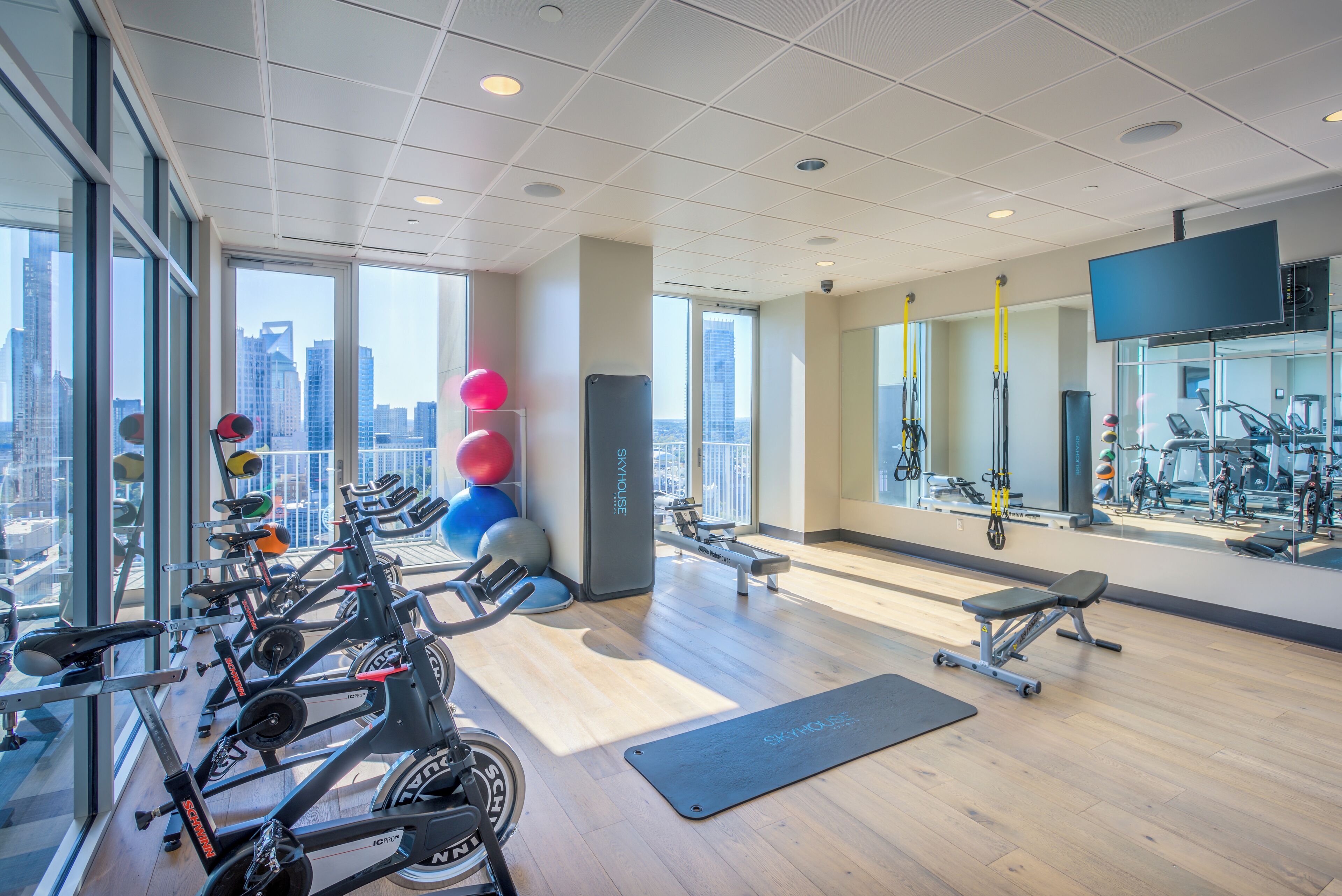 Een moderne sportschool met cardio-apparatuur, gewichten en fitnessballen, grote ramen met uitzicht op de stad en een spiegel die de apparatuur weerkaatst.