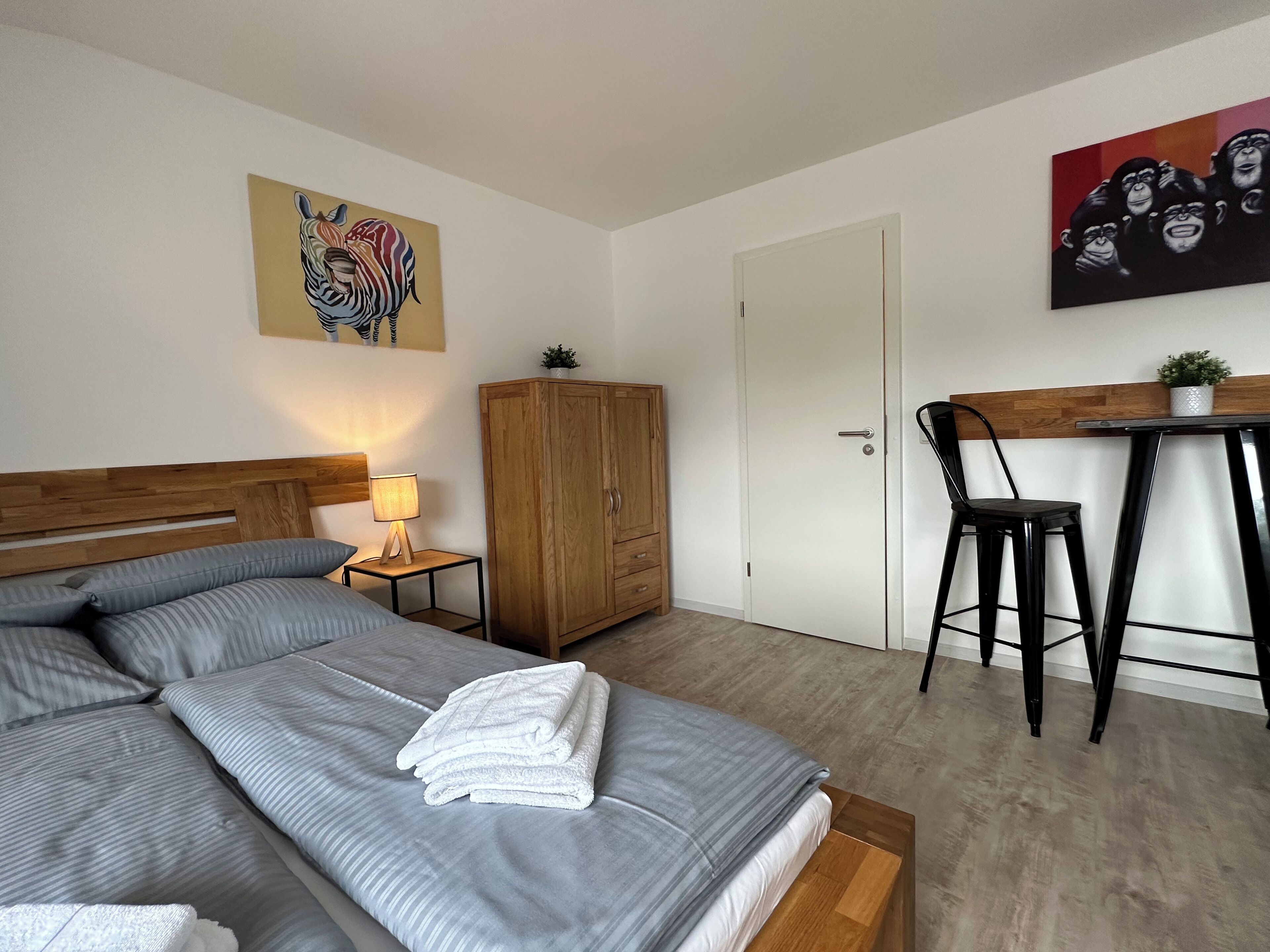 Een keurig opgeruimde slaapkamer met een bed, een houten garderobe, een bureau met stoel en twee schilderijen aan de muur.