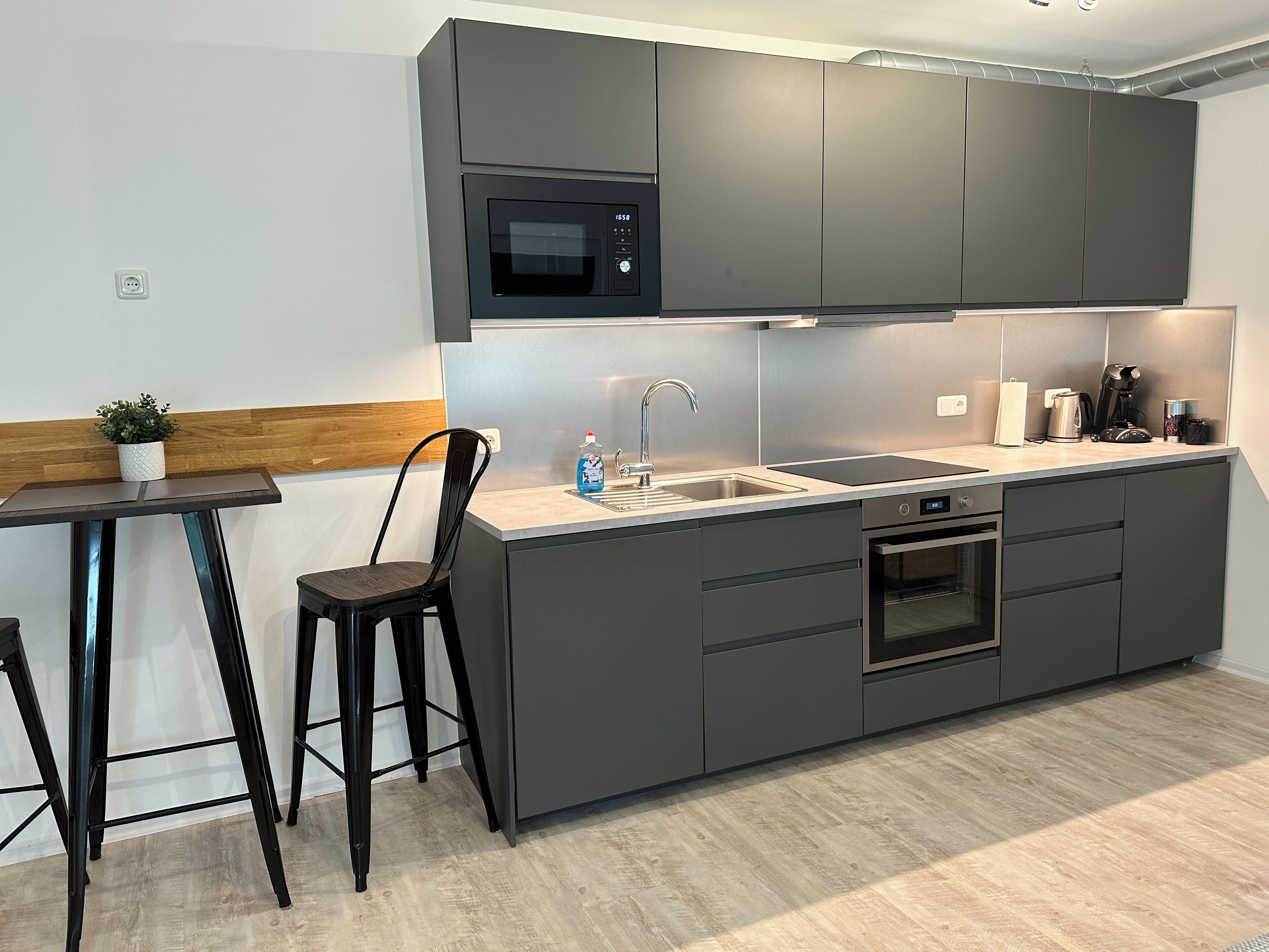 Una cucina moderna con mobili grigi, un forno a microonde integrato, un lavello e un forno. È presente un piccolo tavolo con una sedia e una pianta in vaso.