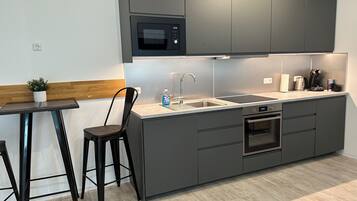 Una cucina moderna con mobili grigi, un forno a microonde integrato, un lavello e un forno. È presente un piccolo tavolo con una sedia e una pianta in vaso.