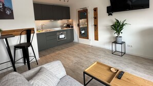 Een moderne keuken met donkere kastjes, een inbouwoven en een zithoek met een grijze bank en een houten tafel.