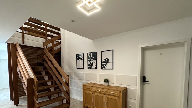 Een trap met houten leuningen die uitkomt op een plateau, een houten kast met lades, en drie ingelijste kunstwerken aan de muur.