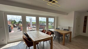 Een ruime kamer met een grote houten eettafel, diverse stoelen en uitzicht op een dakterras door grote ramen.