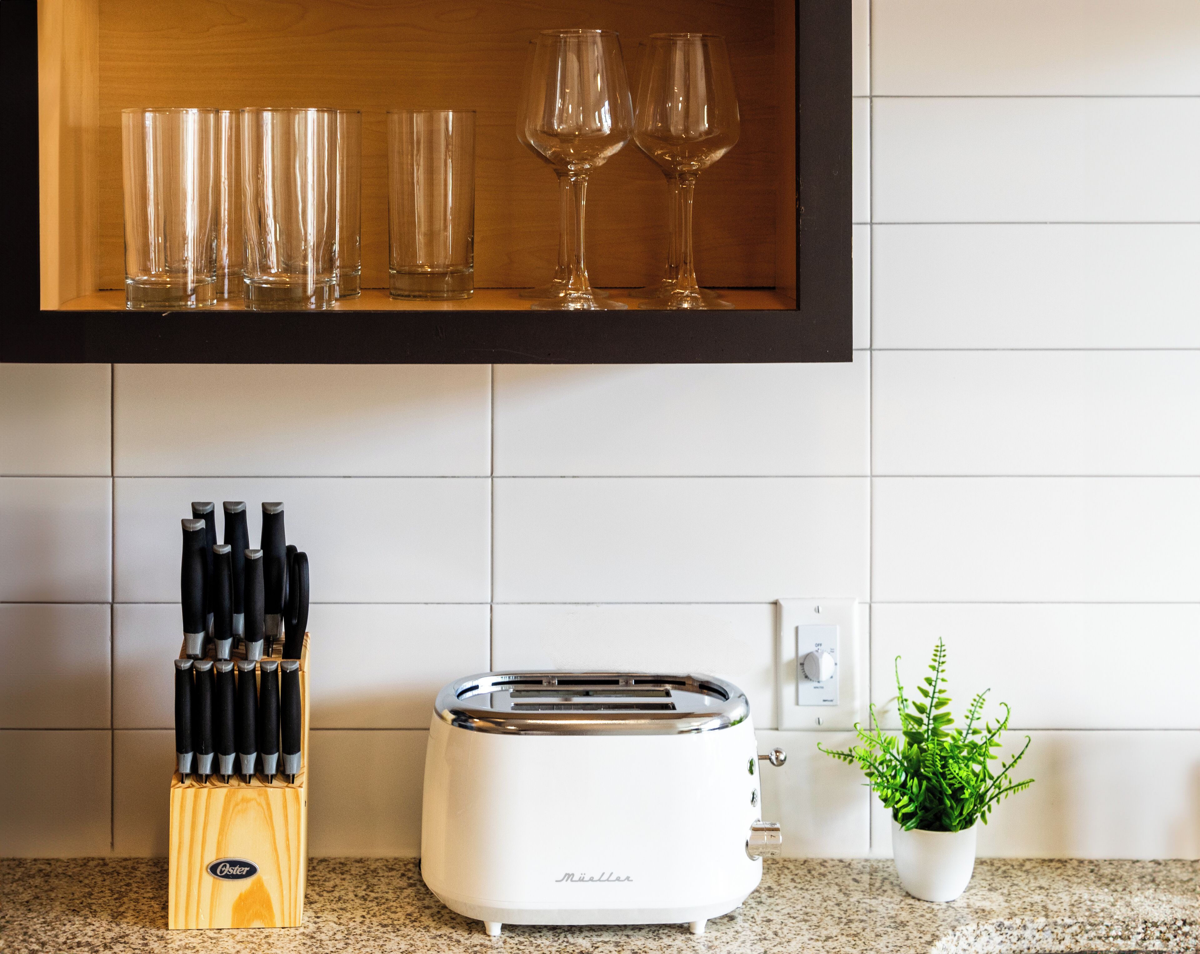 A kitchen with a toaster, a knife block, a potted plant, and a shelf with wine glasses.