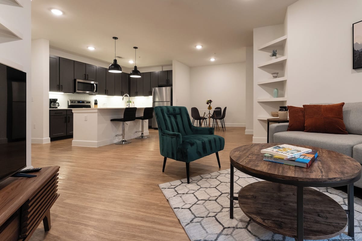 A modern living room with a grey sofa, teal armchair, and a coffee table with books. A kitchen with dark cabinets and a dining area with a table and chairs are visible in the background.