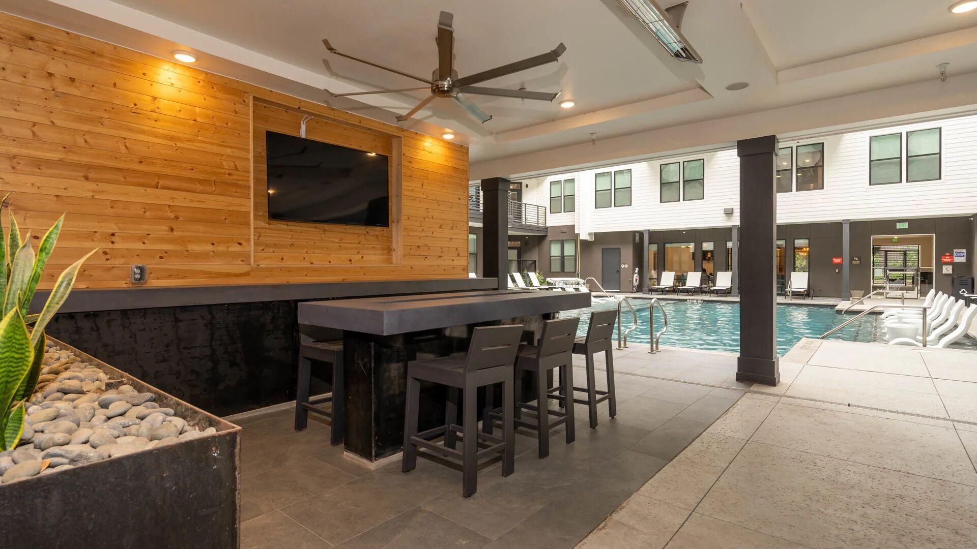 A modern indoor pool area with a bar and seating, wooden paneling, and a ceiling fan.