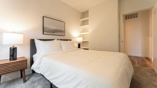 Una cama bien hecha con sábanas blancas, una mesita de noche con lámpara y un cuadro en la pared.