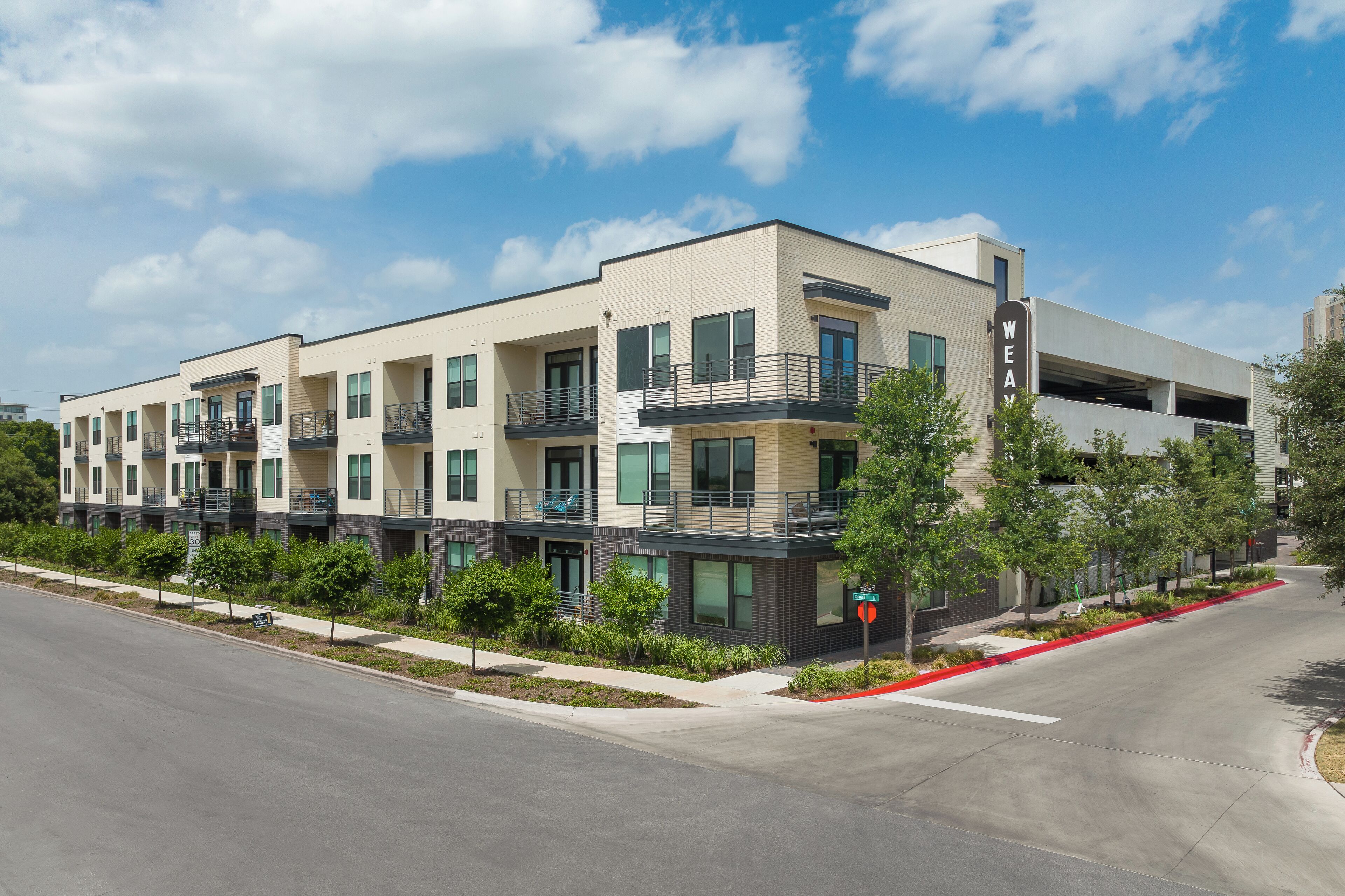 A modern multi-story building with balconies, a parking garage, and a landscaped area with trees.