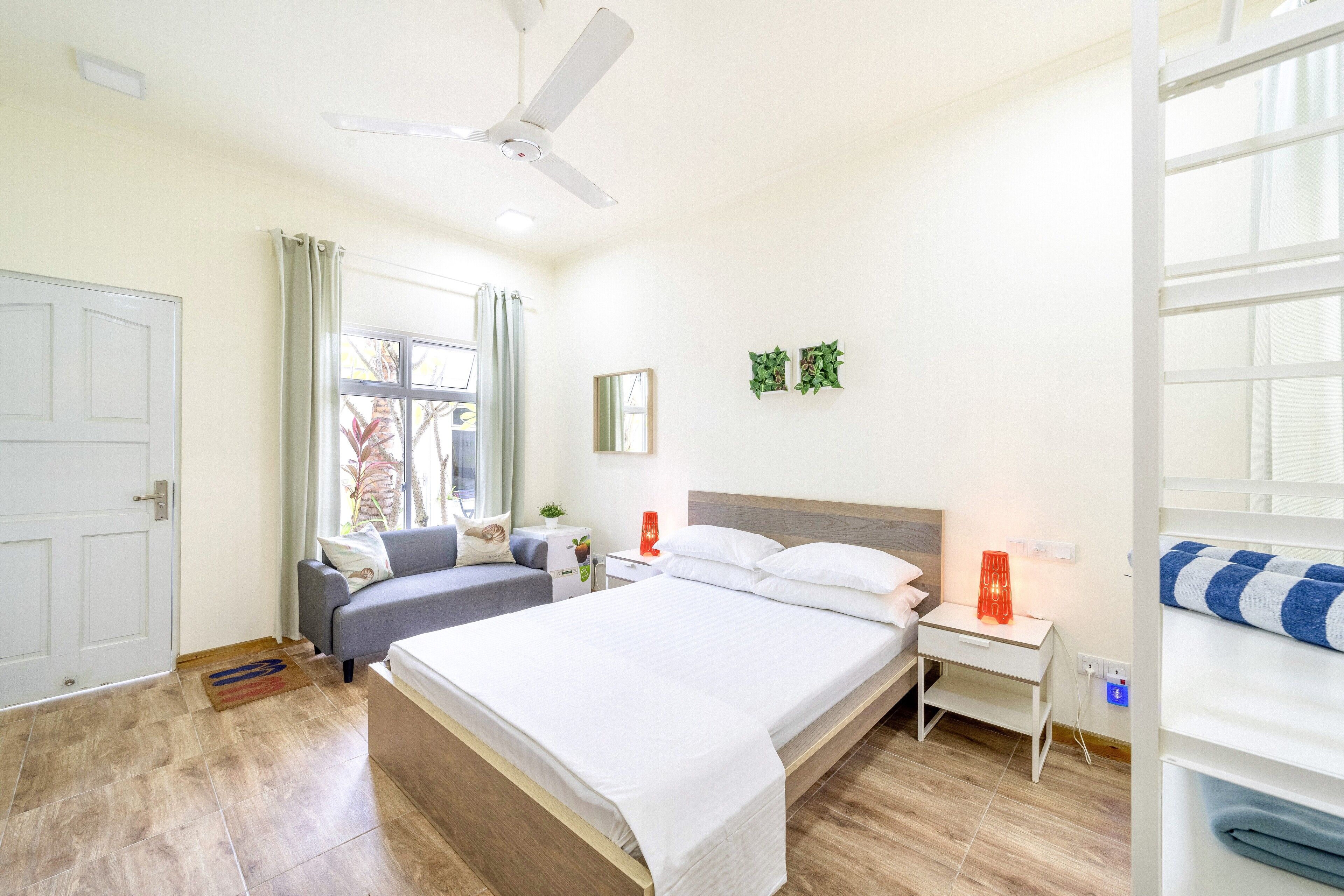 A hotel room with a bed, a desk, a chair, a ceiling fan, a window with curtains, and a door.