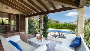Un patio couvert avec des canapés blancs, une petite table sur laquelle se trouve un saladier rempli de fruits et des verres, et une vue sur une piscine et la verdure environnante.
