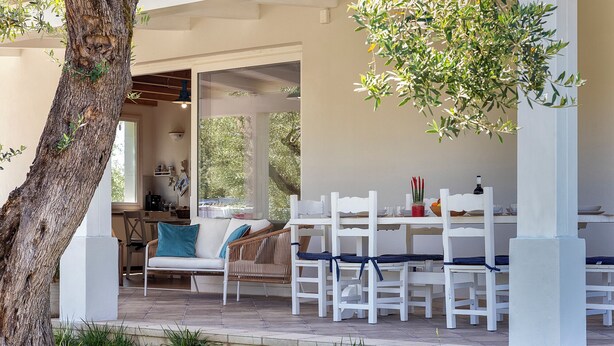 A covered outdoor dining area with white furniture, a tree, and a view of the interior. - ClickSardegna Villa Celeste: Countryside Deluxe near the beaches of Sardinia (Alghero)