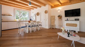 A spacious living area with a wooden ceiling, a dining table with chairs, a kitchen area with a window, and a television on a white cabinet. - ClickSardegna Villa Celeste: Countryside Deluxe near the beaches of Sardinia (Alghero)