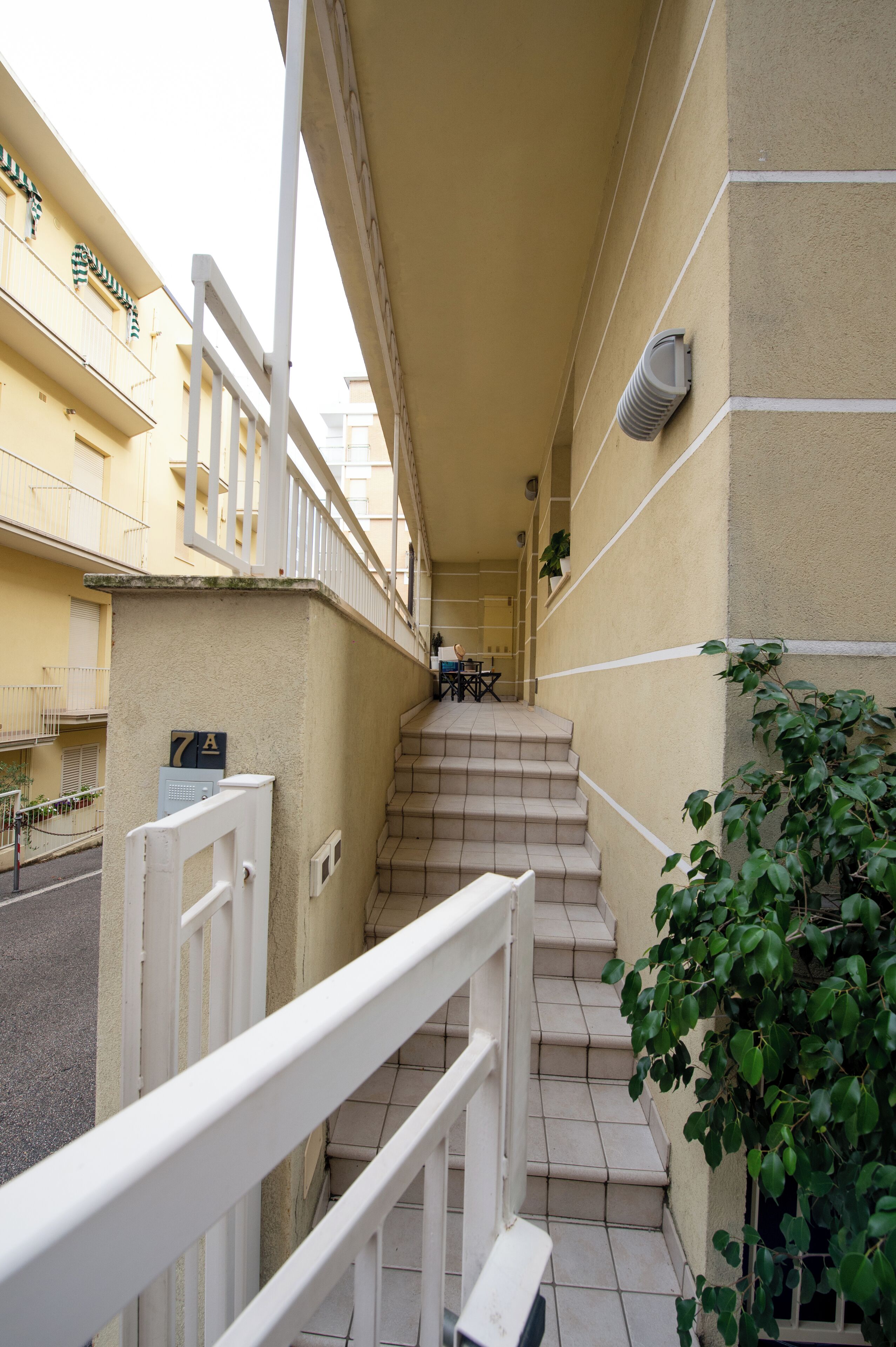 A narrow outdoor corridor with a white railing, beige walls, and a staircase leading to an upper floor.