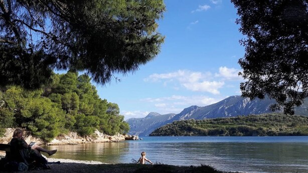 Eine Uferlandschaft mit einer Person, die auf einer Bank sitzt, und einer weiteren Person am Wasser steht.