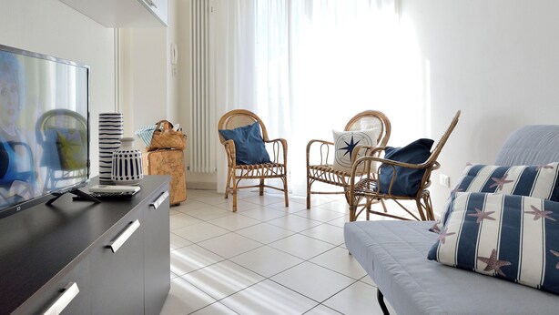 Een moderne woonkamer met een flatscreen-televisie, rieten stoelen, een grijze bank en een betegelde vloer.