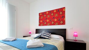Een opgemaakt bed met een blauw-witte deken, twee kussens, en een wanddecoratie met een rood patroon erboven.