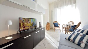 Een moderne woonkamer met een flatscreen televisie, een donker houten kast, een witte bank, rieten fauteuils en een groot raam met dunne gordijnen.