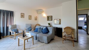Un salon comprenant un canapé, deux fauteuils, une table basse et un ordinateur portable. Les murs sont ornés de tableaux encadrés, il y a un climatiseur fixé au mur et une fenêtre avec des rideaux.