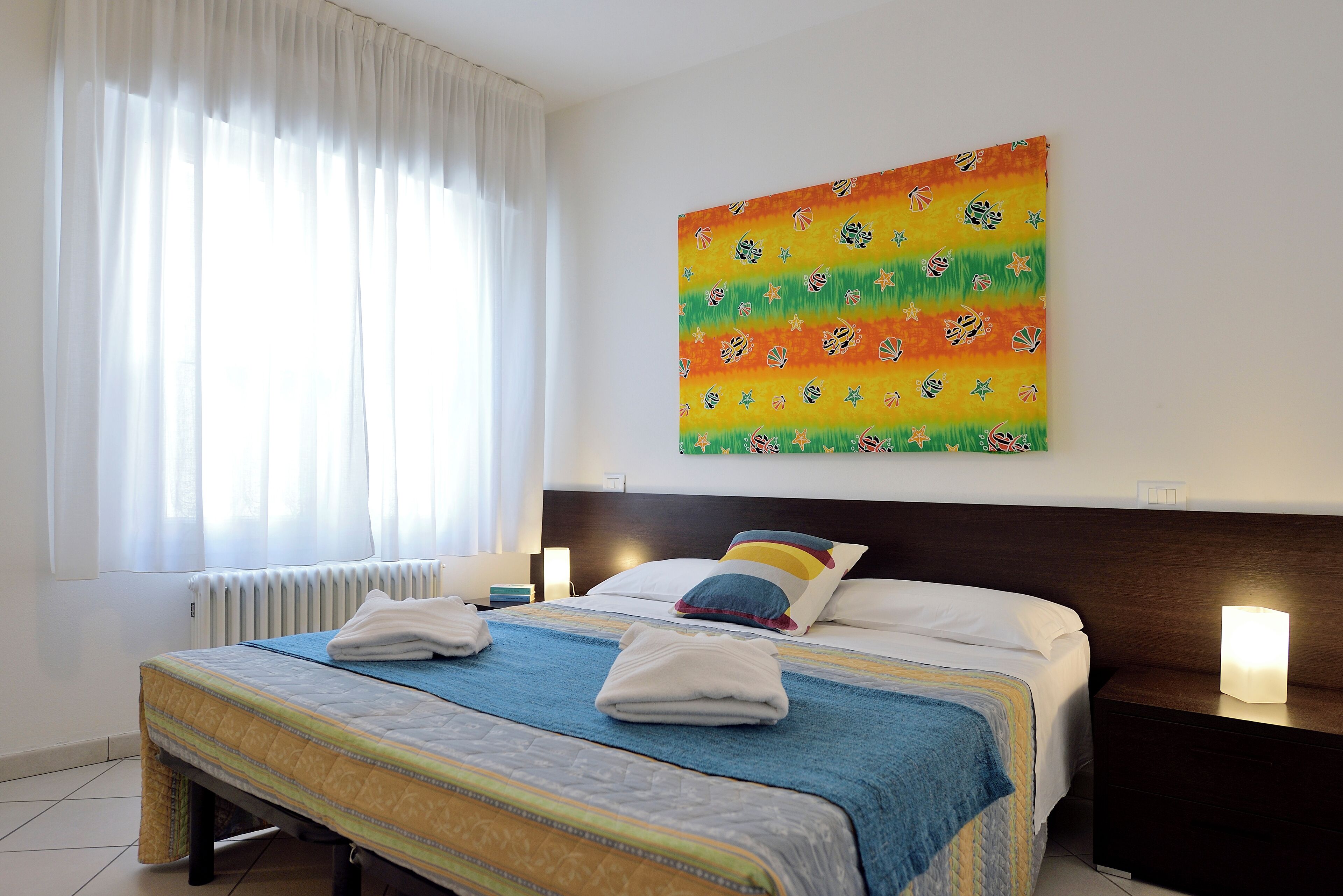A hotel room with a bed, a nightstand, a painting on the wall, and a window with curtains.