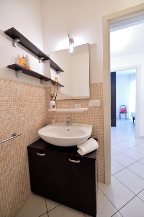 Une salle de bain avec un lavabo blanc, un meuble sombre et des murs carrelés. On trouve des étagères au-dessus du lavabo où sont rangés les articles de toilette, ainsi qu'un miroir au-dessus.