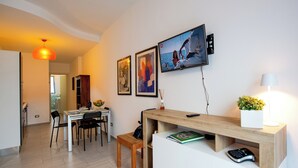 Uma sala de estar moderna com área de refeições, uma escrivaninha de madeira com abajur e uma TV fixada na parede.