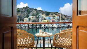 A balcony with wicker chairs, a small round table with a book and a cup, and a view of colorful buildings by the water. - House in Symi with sea view (Simi)