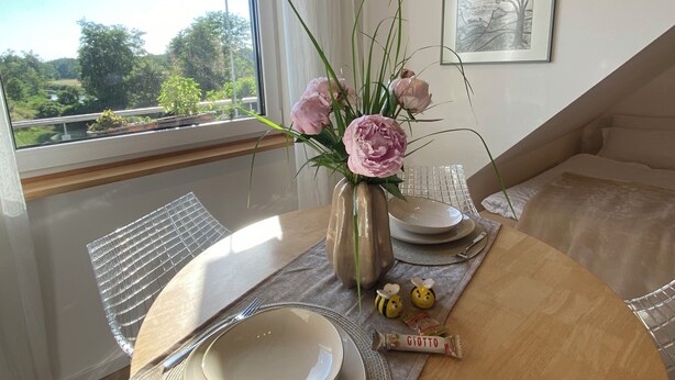 Een eethoek met een tafel gedekt voor twee personen, een vaas met bloemen en uitzicht op groen.