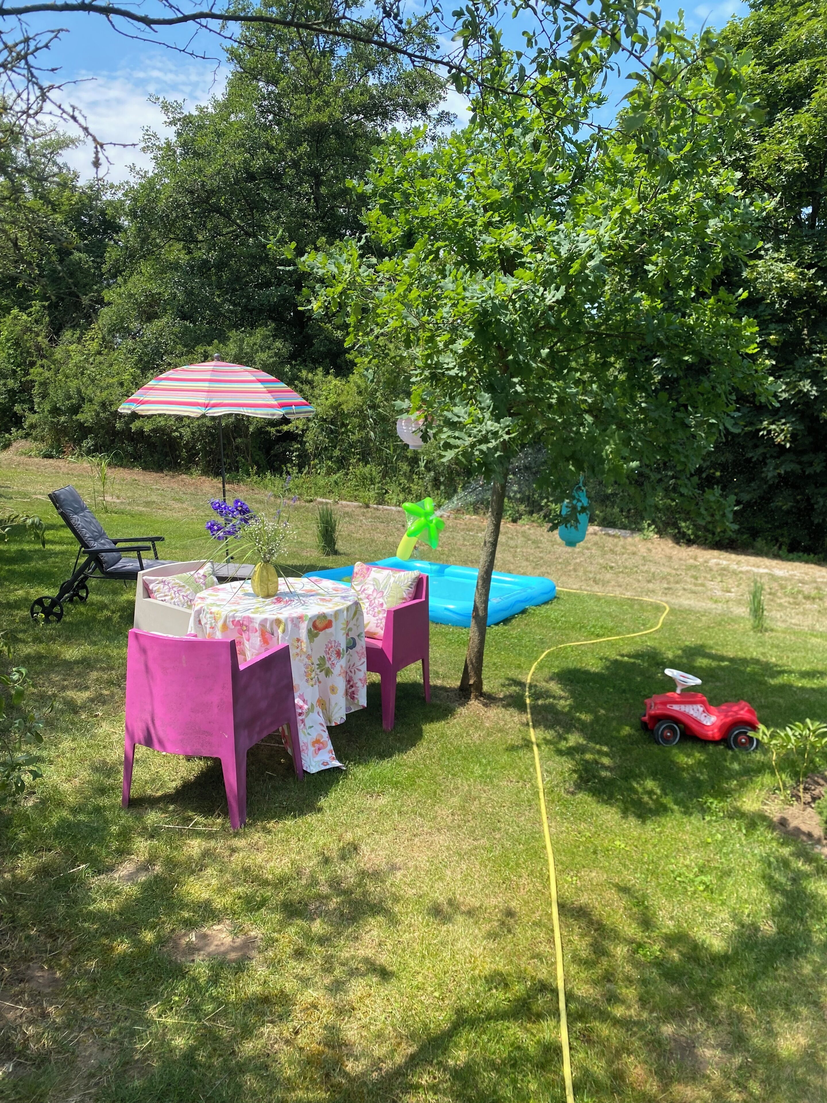 A backyard with a table, chairs, a colorful umbrella, a small inflatable pool, and a toy car.