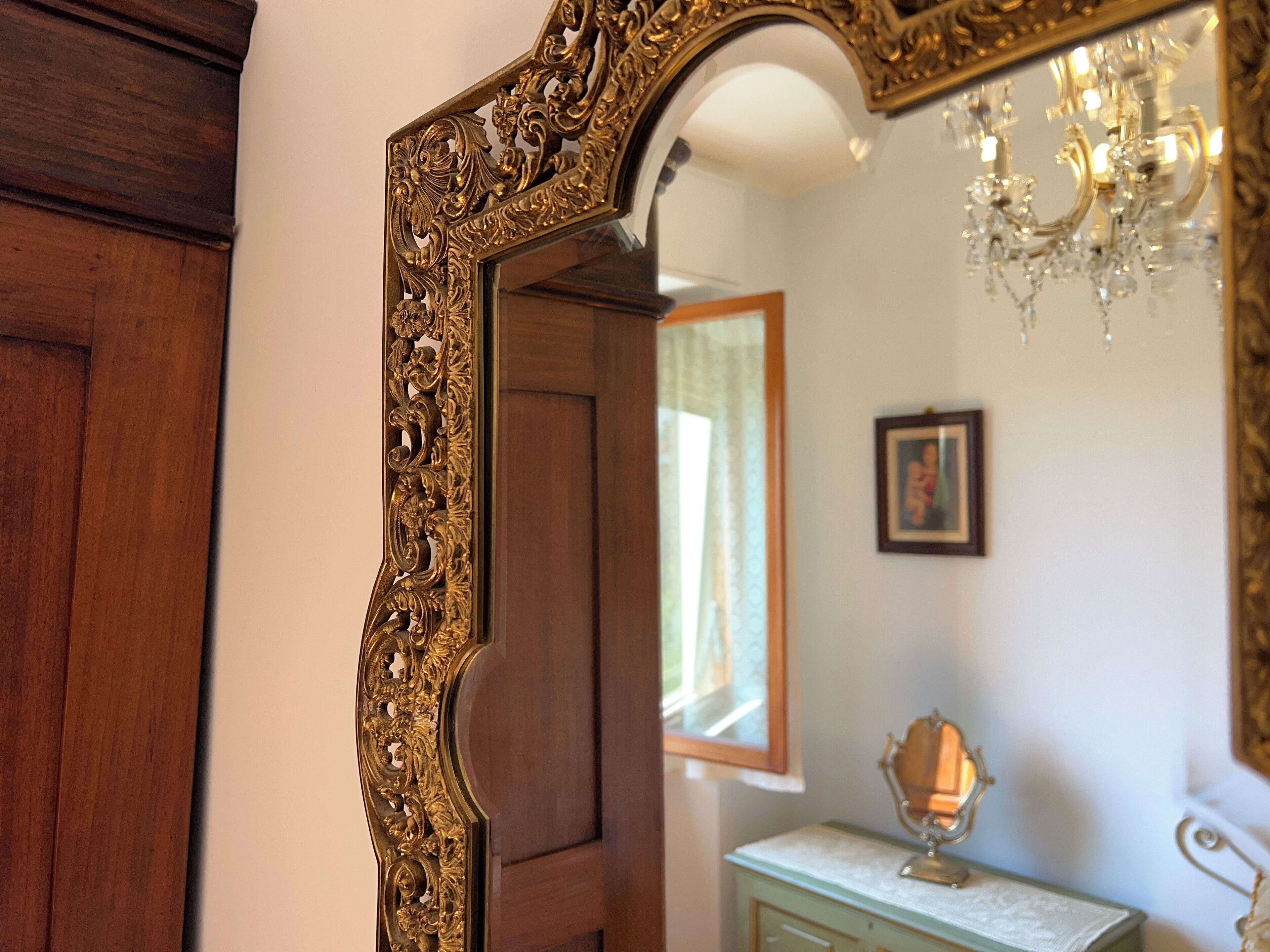 Ein verziertes Spiegelbild mit goldener Rahmung, eine Holztür, ein Fenster mit Vorhängen, ein kleiner Tisch mit Lampe und ein an der Wand befestigtes Bild.