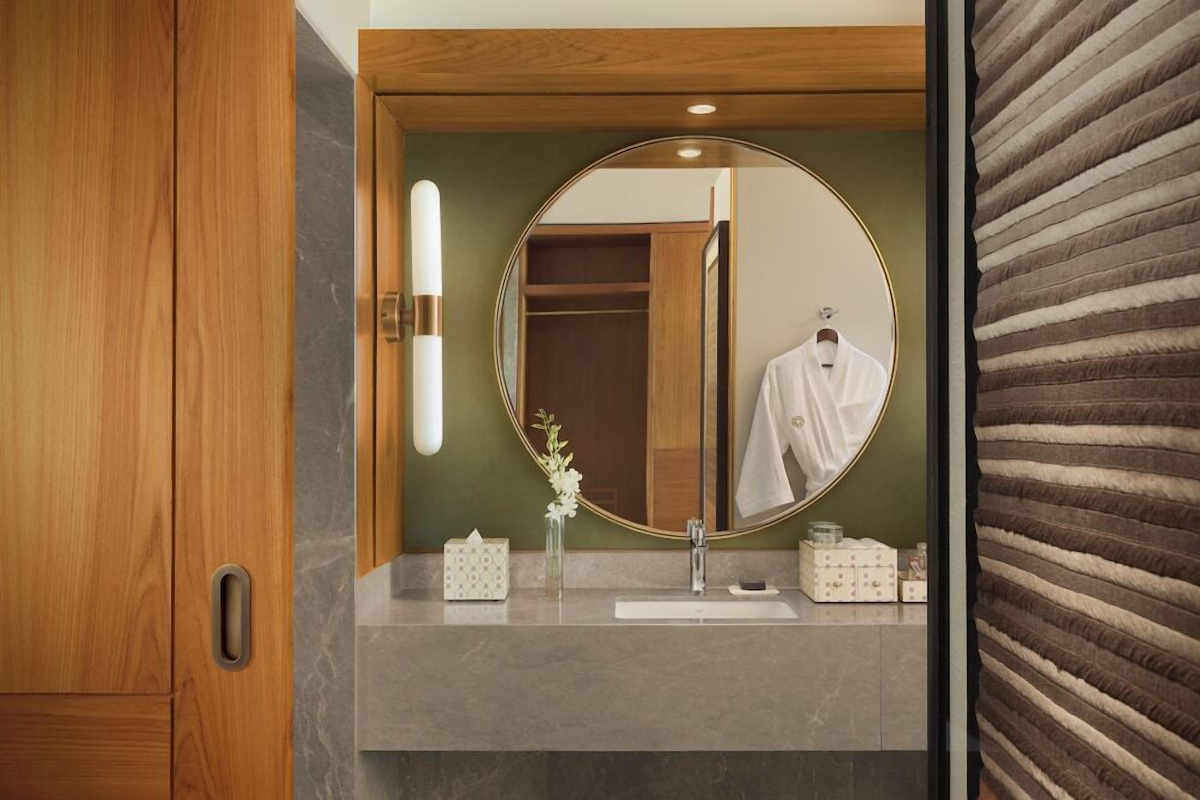 Un baño con puerta de madera, un espejo redondo, una toalla blanca, un florero con flores y un lavabo con artículos de tocador.
