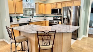 A kitchen with wooden cabinets, a stainless steel refrigerator, a microwave, and a stove. There are two bar stools and a clock on the wall. - Beautiful Bungalow - 3/2 w/ Golf Cart in The Village of Liberty Park SMALL DOG Accepted - Max 1x 20-25lbs (The Villages)