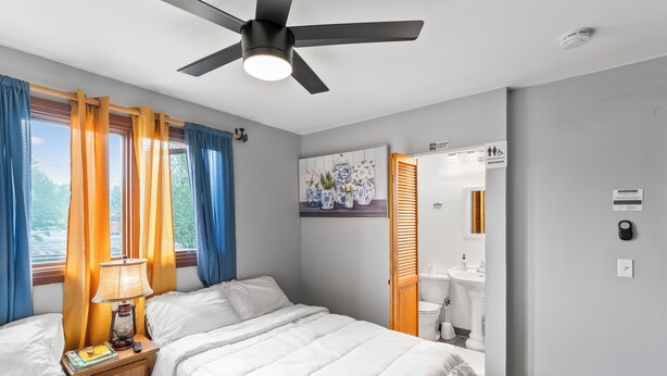 Une chambre à coucher avec un lit, une table de chevet, une lampe et des rideaux. On y trouve également un ventilateur de plafond et une fenêtre avec des rideaux.