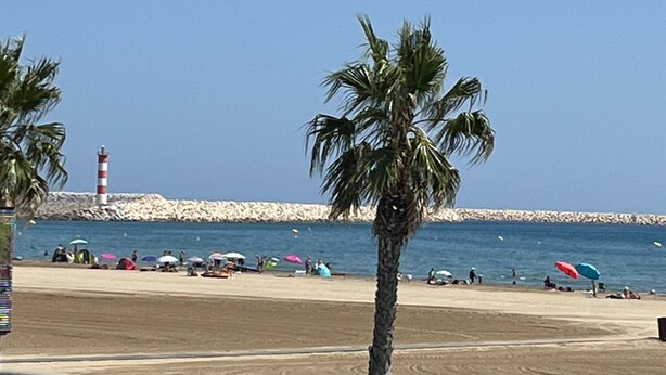 Ein Strand mit Palmen, einem Leuchtturm und Menschen unter Sonnenschirmen.