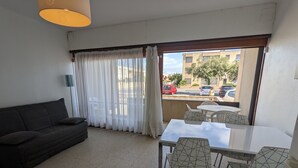 Ein Wohnzimmer mit einer Couch, einem Esstisch mit Stühlen, einem Fenster mit Vorhängen und Blick auf die Straße.