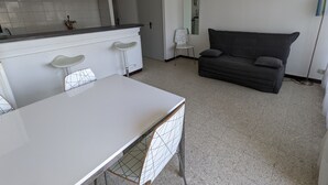 Eine moderne Küche mit weißen Schränken, einer weißen Arbeitsplatte und einem Esstisch mit einem Stuhl mit geometrischem Muster. Im Wohnbereich befindet sich eine dunkelgraue Couch und eine Stehlampe.