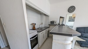 A compact kitchen with a microwave, oven, and a sink. There is a clock on the wall and a sofa in the adjacent living area. - Seafront T2 with AC and parking (Port-la-Nouvelle)