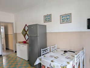 A kitchen with a refrigerator, a dining table with chairs, a small cabinet, and a wall-mounted picture frame.