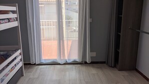 A room with a wooden floor, a sliding glass door with white curtains, a wooden wardrobe, and a green hanger on the wall. - Charming 2-Bedroom Villa with AC, 400m from Beach, Sleeps 5/6 (Port-la-Nouvelle)