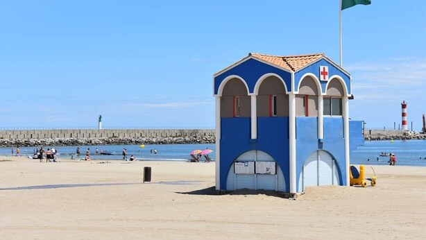 A beach lifeguard tower with a red cross, a flag, and a sandy beach with people and a pier in the background. - Charming 2-Bedroom Villa with AC, 400m from Beach, Sleeps 5/6 (Port-la-Nouvelle)