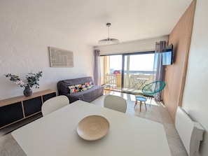 A modern living room with a sofa, a dining table with a bowl, a balcony with chairs, and a view of the sea. - Seafront 1BR Apartment with Balcony, Parking & Stunning Views - Sleeps 5 (Port-la-Nouvelle)