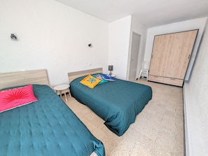 A bedroom with two beds, a small table, a lamp, and a wardrobe. - Seafront 1BR Apartment with Balcony, Parking & Stunning Views - Sleeps 5 (Port-la-Nouvelle)