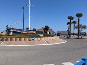 A roundabout with a blue directional sign, a colorful boat sculpture, palm trees, and a building with a red roof in the background.