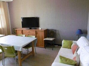 Een kamer met een eettafel, stoelen, een televisie op een houten kast, een bank met kussens, en een lamp.