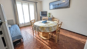 A dining area with a round table, chairs, a ceiling fan, a television, and a window with curtains. - Charming Ground-Floor T2 Apartment, 300m from Beach, Port View, Parking Included, Sleeps 4. (Port-la-Nouvelle)