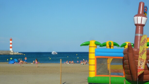 A beach with a large inflatable play structure, a lighthouse, and a boat in the background. - Charming Ground-Floor T2 Apartment, 300m from Beach, Port View, Parking Included, Sleeps 4. (Port-la-Nouvelle)