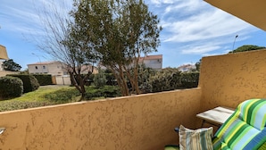 A balcony with a striped sofa and a small table, overlooking a residential area with houses and greenery. - Charming Ground-Floor T2 Apartment, 300m from Beach, Port View, Parking Included, Sleeps 4. (Port-la-Nouvelle)