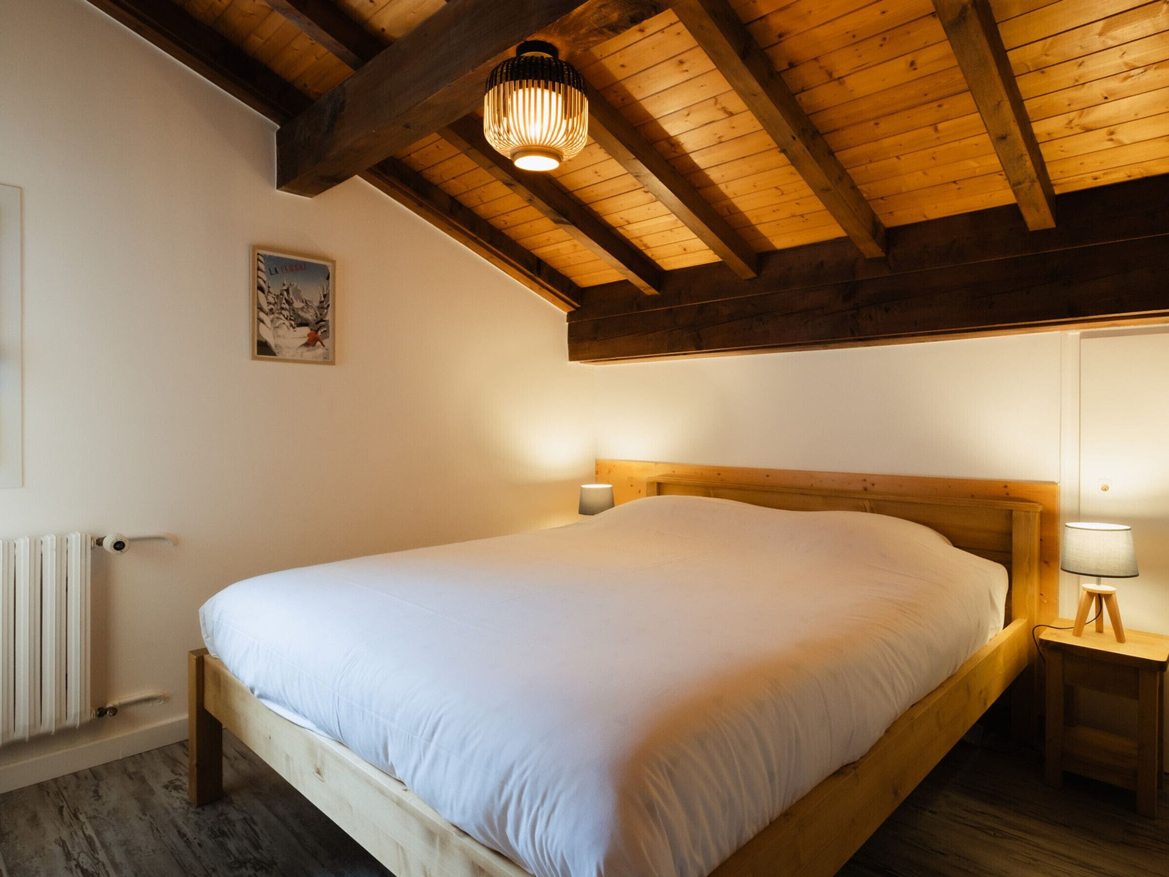 Un dormitorio con una cama de madera, una mesita de noche con lámpara, un radiador y techo de madera.