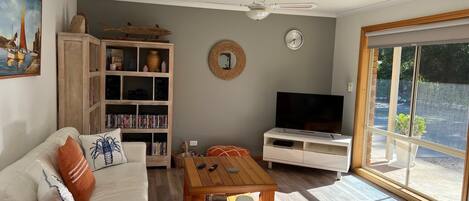 Una sala de estar con un sofá blanco, una mesa de centro, una estantería, un televisor y un ventanal grande.
