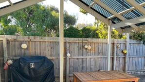 A covered patio with a wooden table and bench, a barbecue grill, and a wooden fence with hanging ornaments.