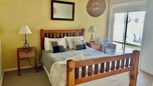 A bedroom with a wooden bed, two bedside tables with lamps, a wicker basket, a mirror, and a view of the outside through a large window. - Lazy Sails Starboard  - Pets welcome outside, WiFi (Robe)
